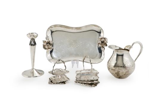 150   -  <p><span class="object_title">Lote en plata española punzonada compuesto por: candelero, jarrita para vino con decoración cincelada de uvas y hojas de parra, dos juegos de ceniceritos en forma de cesta con decoración de flores y bandeja tarjetero con asas a modo de rosas y centro con iniciales grabadas. </span><br></p>