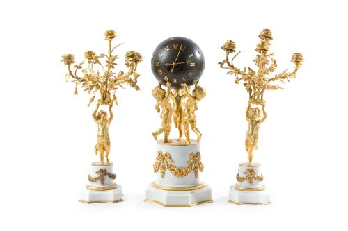 908   -  <p><span class="object_title">Guarnición de chimenea formado por reloj y pareja de candelabros estilo Luis XVI, Francia, s. XIX<br/>En bronce dorado y mármol blanco. <br/>Reloj con esfera sostenida por tres putti sobre pedestales con guirnaldas. Numeración romana y agujas en forma de flechas. Maquinaria tipo París.<br/>Pareja de candelabros a juego, fuste a modo de putto que sostiene 5 brazos de luz rameados. Sobre base circular con guirnadas de flores y lazos. </span><br></p>