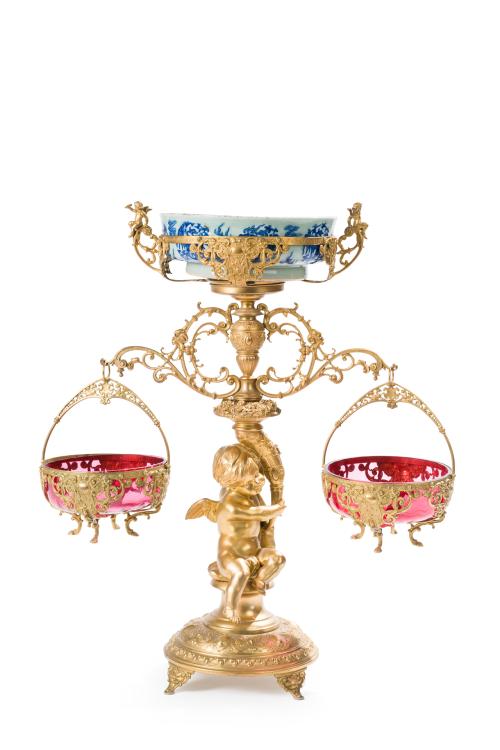 345 - <p><span class="object_title">Decorativo centro de mesa con fuste de angelote portante en metal dorado calado y cincelado, flanqueado por cestas colgantes con recipientes en cristal rojo rubí. Remate con cuenco en porcelana china blanca y azul (consolidado).</span><br></p>