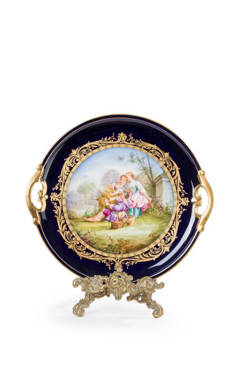 188 - <p><span class="object_title">Bandeja de presentación en porcelana de Sèvres en azul cobalto con escena campestre en el campo orlada con cenefa en dorado, firmada. Con marcas.<br/>Sobre atril en bronce dorado (con faltas). </span><br></p>